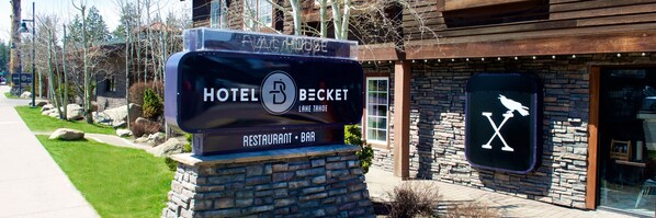 Letrero de hotel con fachada rústica, base de piedra y logotipo que indica que ofrece restaurante y bar.