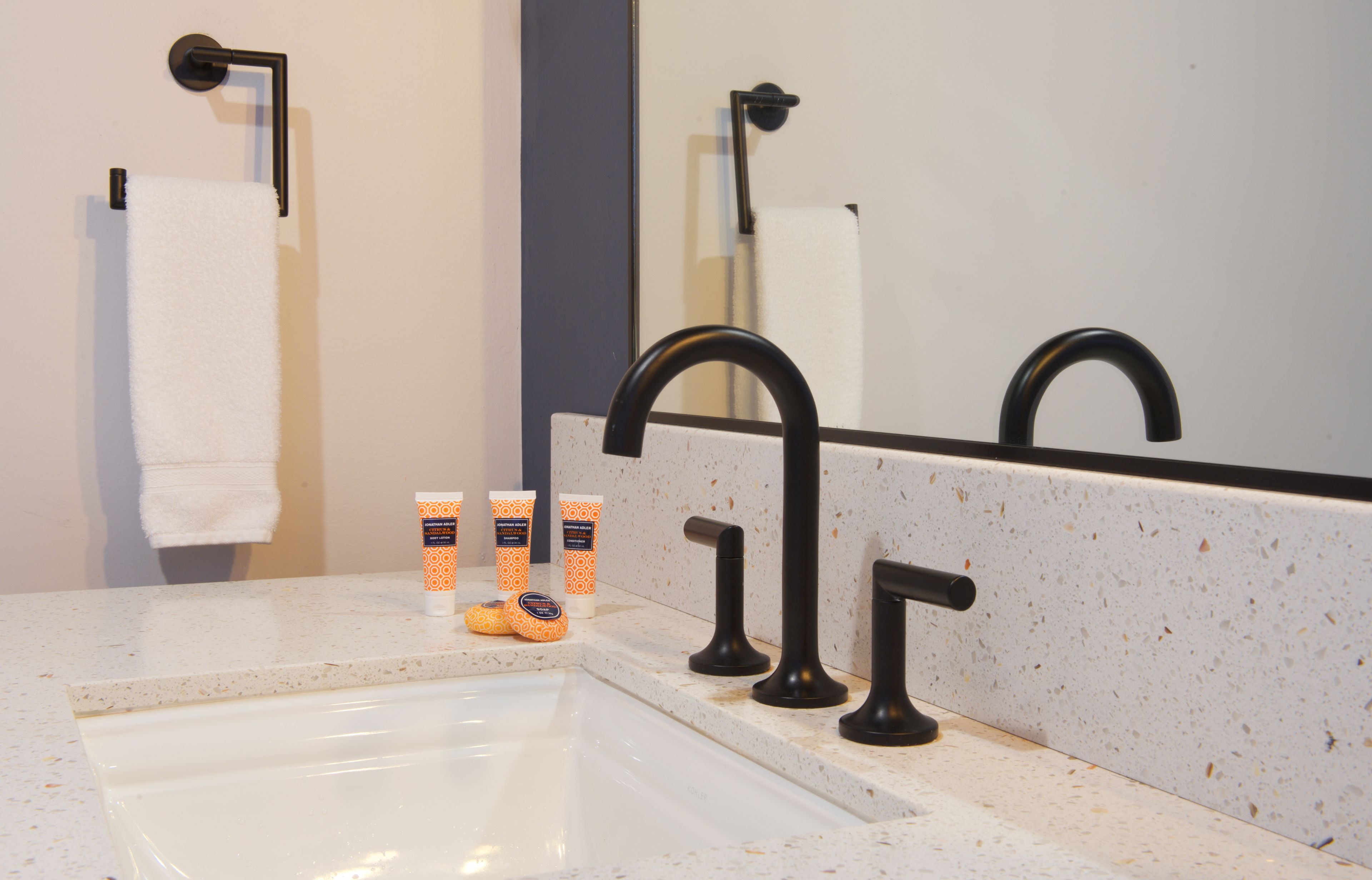 Een badkamer met een wit wastafel, zwarte kraanwerk en een spiegel. Op het aanrecht liggen twee rollen toiletpapier en twee oranje en witte voorraadjes toiletartikelen.