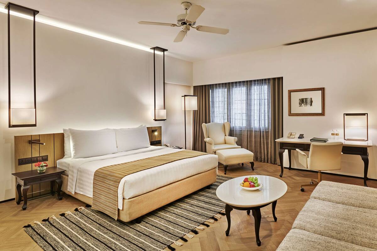 Habitación de hotel con cama, escritorio, silla, una mesita redonda con fruta, lámpara de pie y ventilador de techo.