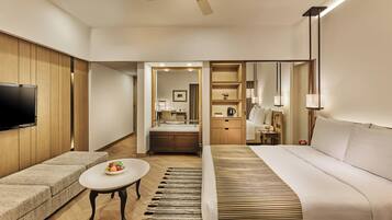 Habitación de hotel con sofá, una mesita con fruta, cama, televisor montado en la pared, ventilador de techo y una zona de kitchenette.