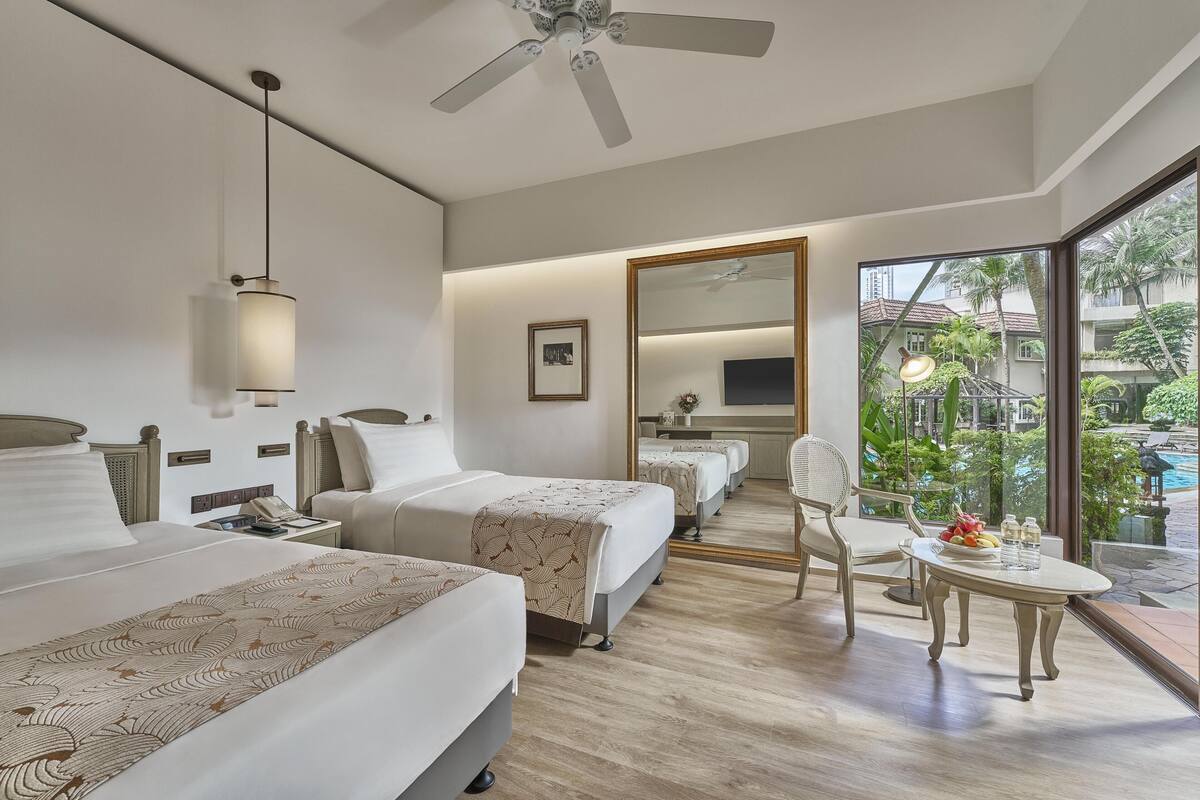Habitación de hotel con dos camas, un escritorio, una silla, una mesita con fruta, un ventilador de techo y un amplio ventanal con vista a una piscina y follaje.