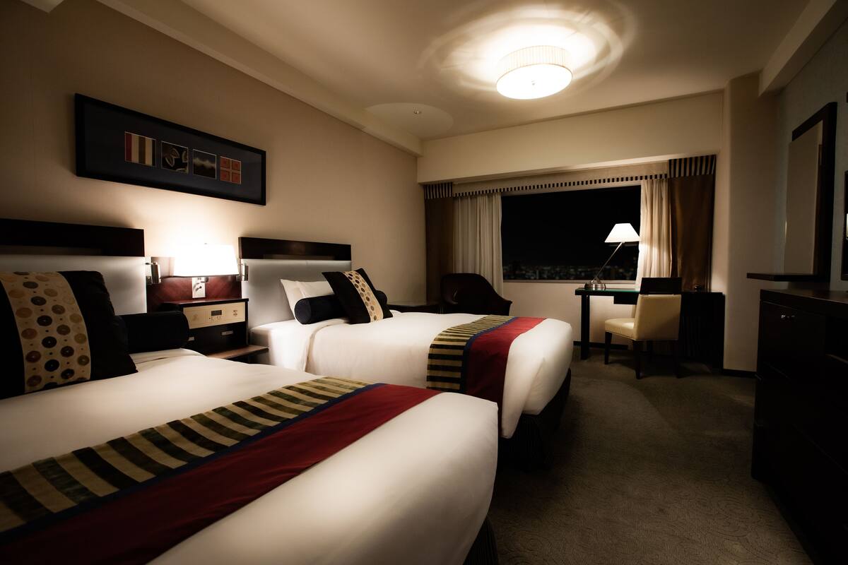 Une chambre d’hôtel avec deux lits, un bureau, une chaise, une lampe et une grande fenêtre avec des rideaux.