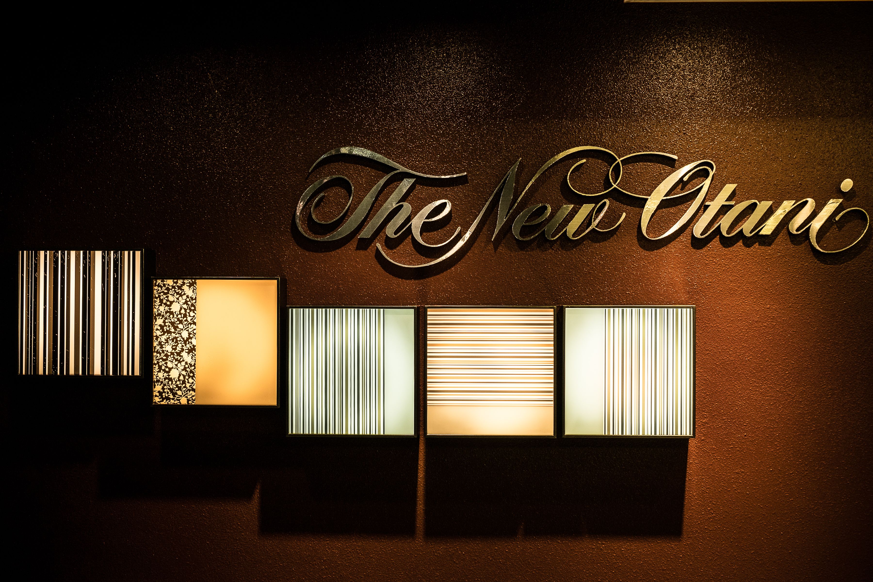 Eine Wand mit beleuchtetem Text "The New Otani" und drei Lichttafeln mit vertikalen Linien.