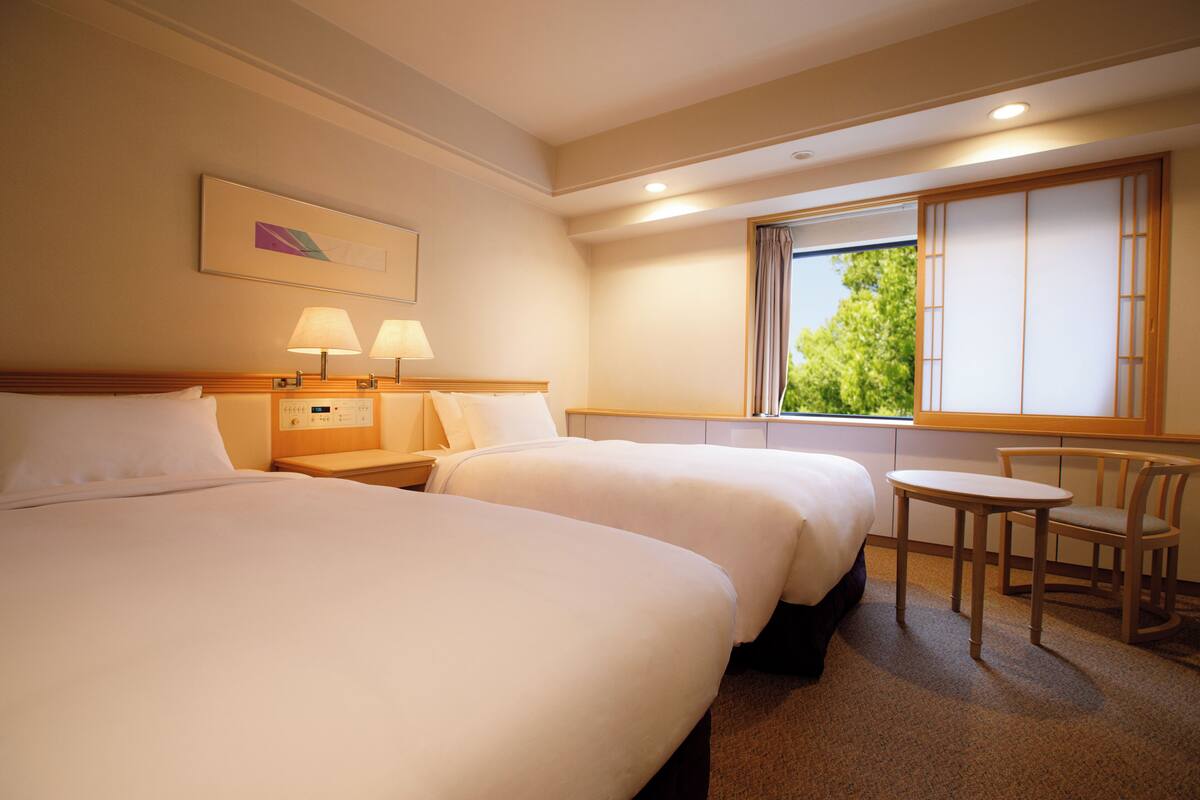 Une chambre d’hôtel avec deux lits, une petite table, une chaise et une fenêtre donnant sur un paysage verdoyant.