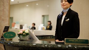 Un comptoir d’accueil d’hôtel avec un panneau indiquant le service aux clients, un vase de fleurs et un écran numérique.