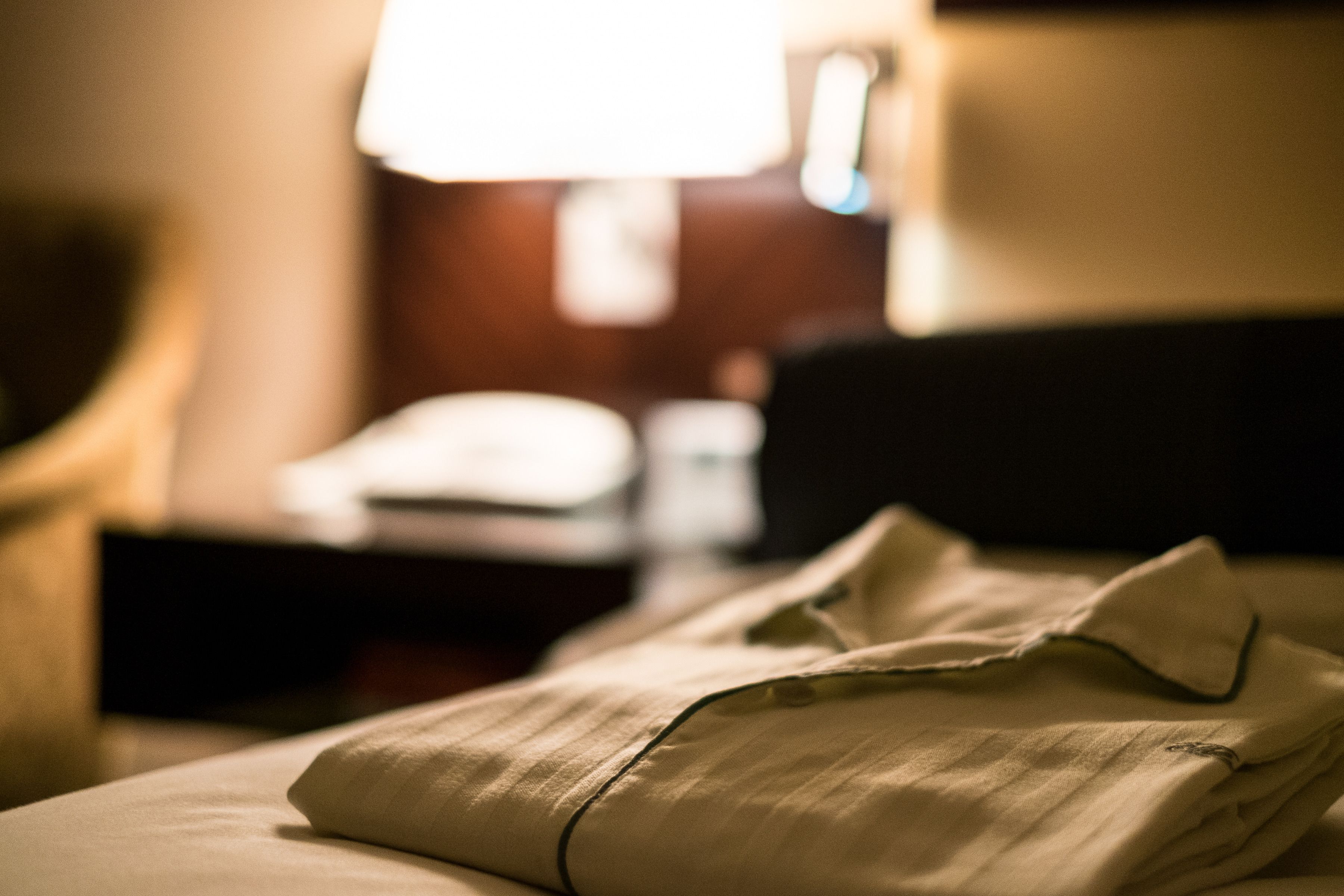 Ein ordentlich gemachtes Bett mit einem gefalteten Hemd, eine Lampe im Hintergrund und ein unscharfer Raum.