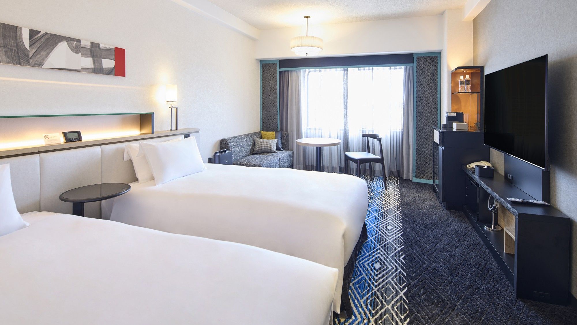 A hotel room with two beds, a TV, a desk, a chair, a small table, and a window with curtains.