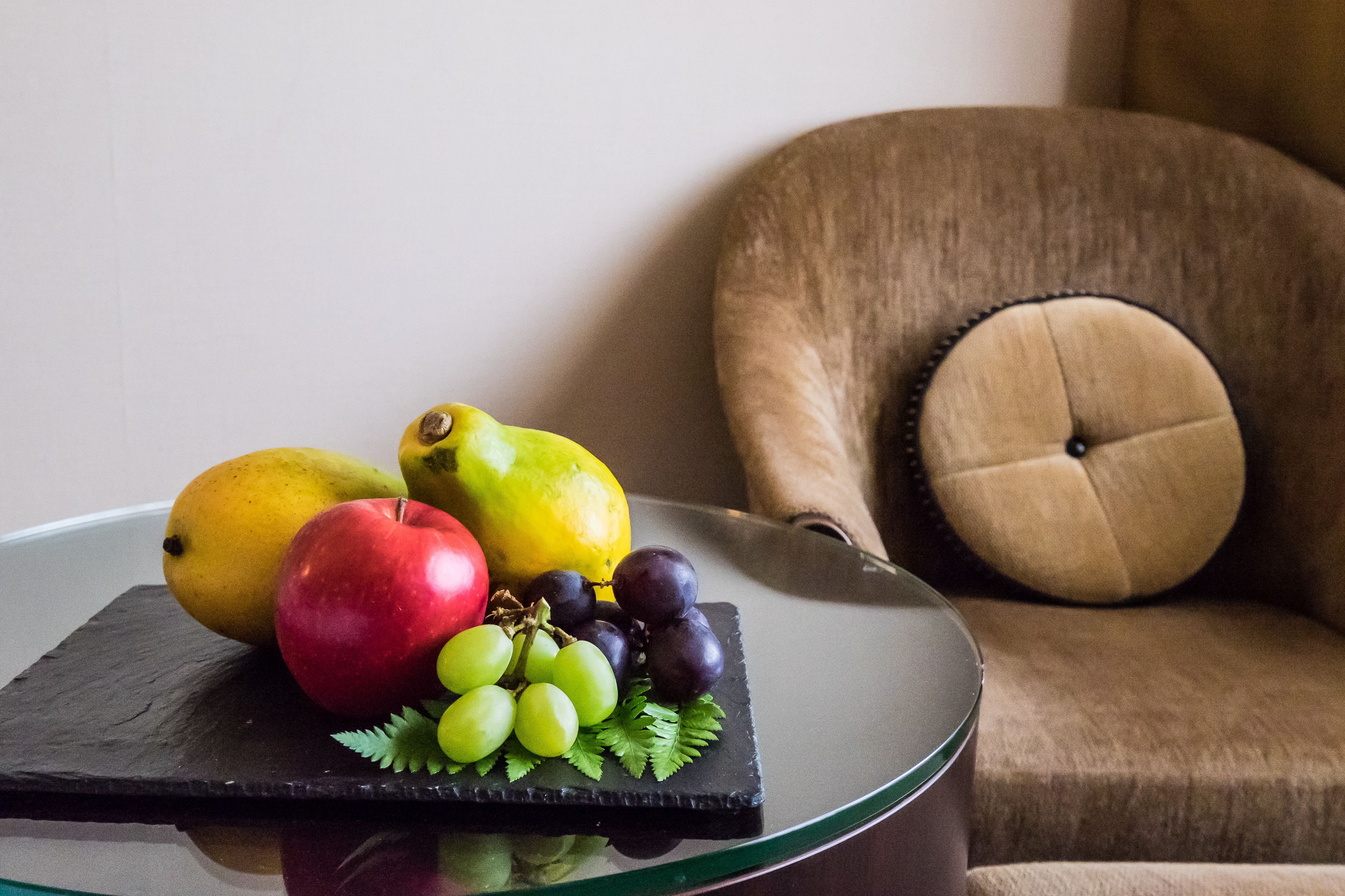 A fruit platter on a glass table with a knife, a brown sofa with a tufted pillow, and a plain wall in the background.