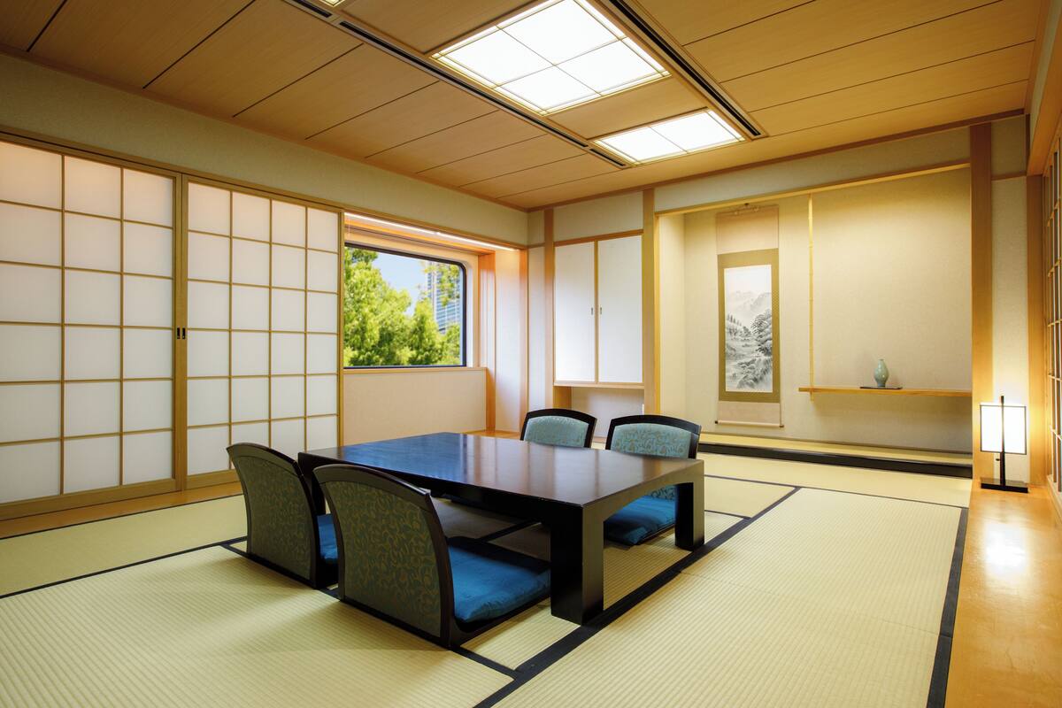 Une pièce traditionnelle avec tatamis, une petite table et des chaises, un paravent en papier de riz (shoji), et une vue sur la verdure à l’extérieur.