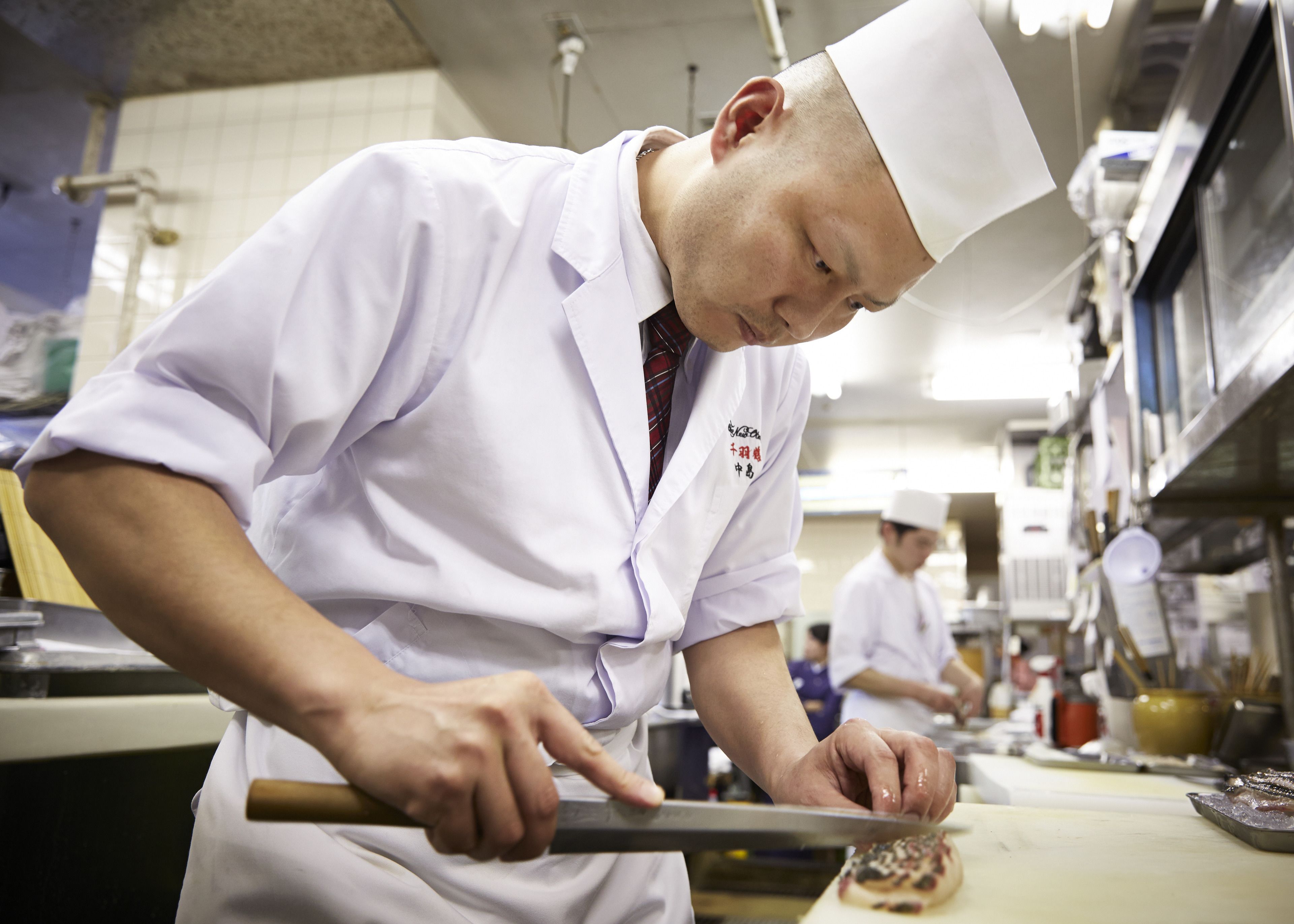 A chef in a white uniform using a knife on a cutting board in a kitchen.