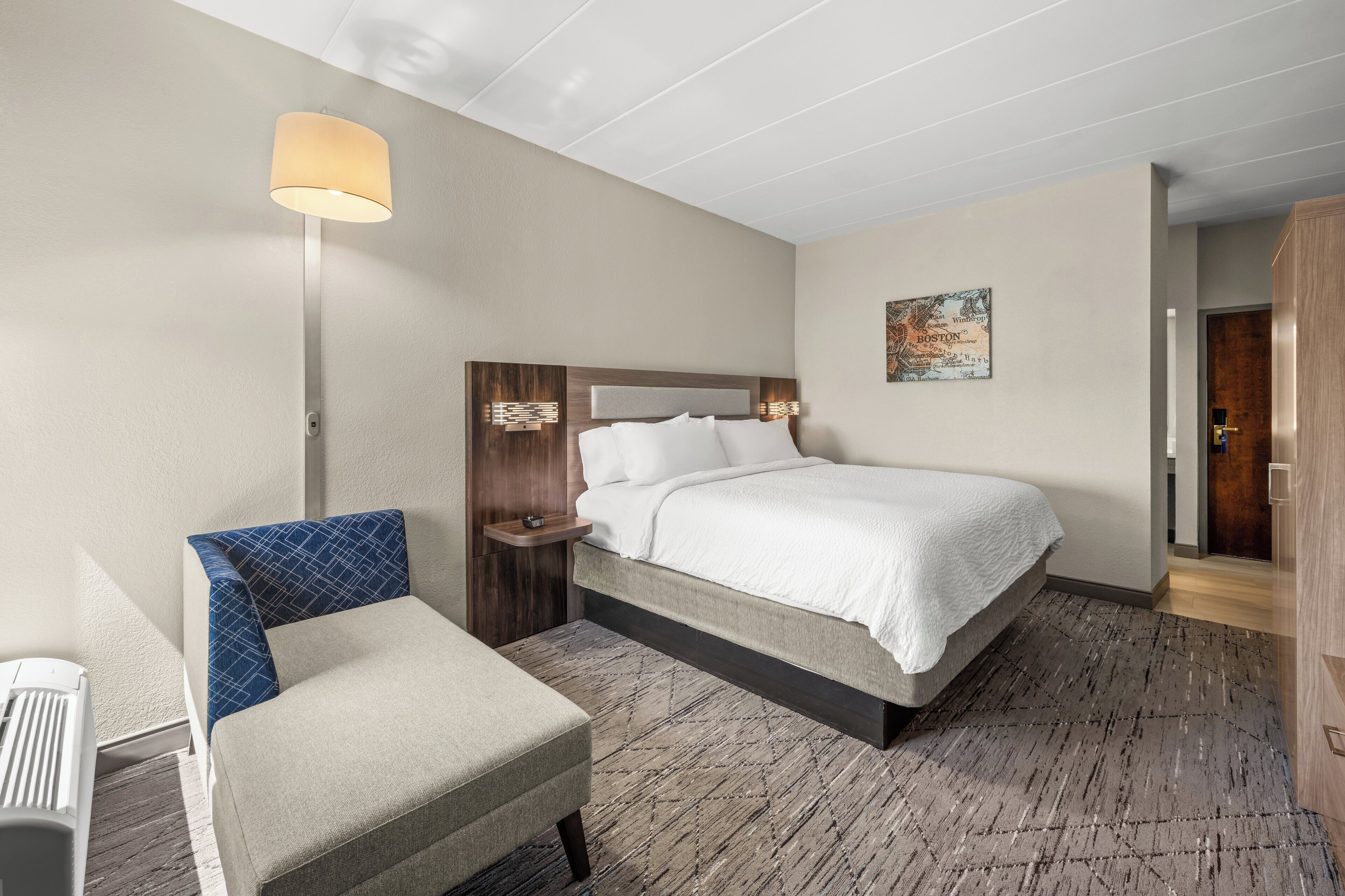A hotel room with a bed, a chair, a desk, and a lamp.