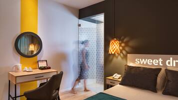 A modern hotel room with a bed, a desk, a chair, a mirror, a shower area, and a nightstand with a phone.