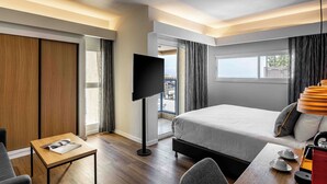 Quarto de hotel moderno com uma cama grande, televisão de tela plana, uma mesinha com uma revista e vista para o exterior.