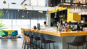 Uma área de bar moderna com balcão de madeira, banquetas pretas e diversas garrafas e copos.