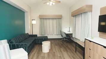 A modern hotel room with a sectional sofa, a desk, a chair, a small ottoman, a ceiling fan, and a TV.
