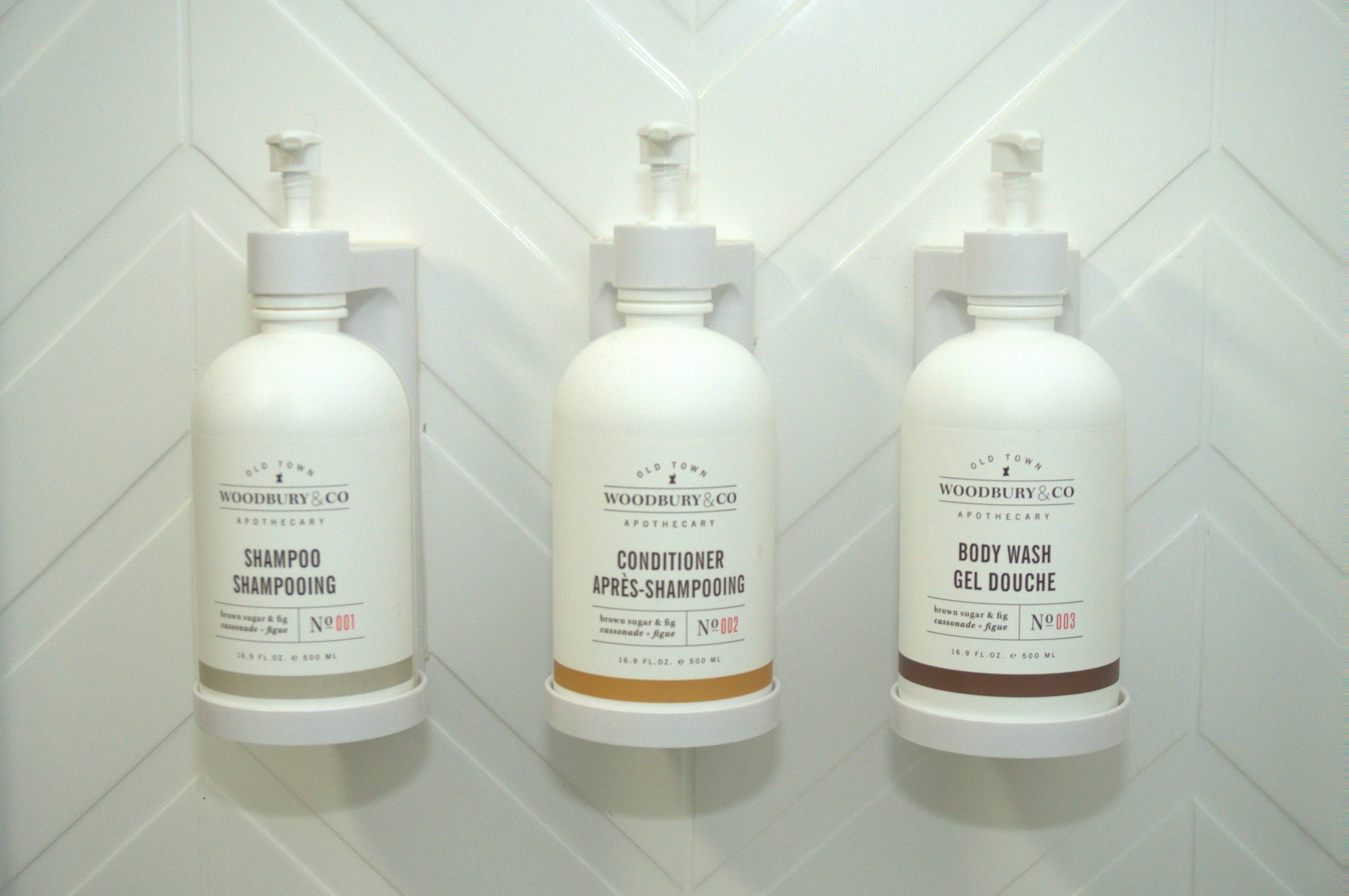 Tre flaskor med produkter från Old Town Woodbury & Co. står på en vit vägg täckt med kakel.