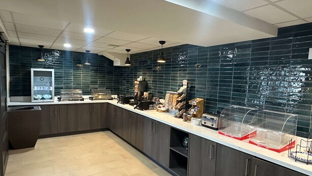A modern kitchen with dark tiled backsplash, wooden cabinets, and a countertop with various appliances and food display cases. - La Quinta Inn by Wyndham Albuquerque Airport (Albuquerque)
