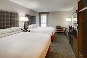 A hotel room with two beds, a desk, a chair, a small table, and a refrigerator.