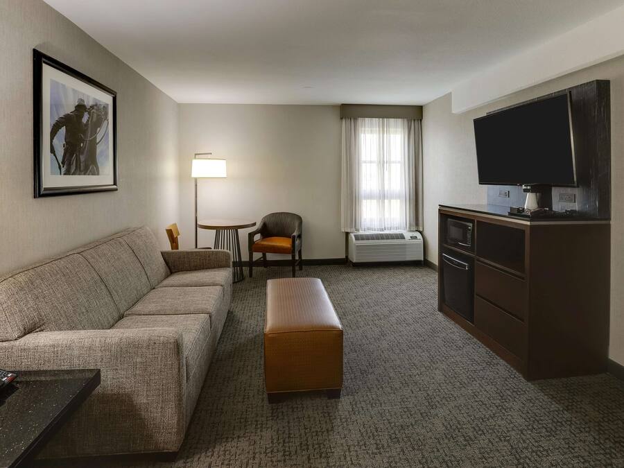 Een hotelkamer met een bank, een kleintje tafeltje met een stoel, een televisie, een kast en een foto in een lijst aan de muur.