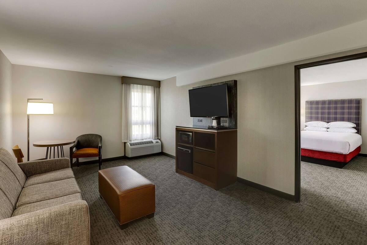 A hotel room with a sofa, a small table, a chair, a TV, a minibar, and a bed with a red accent.