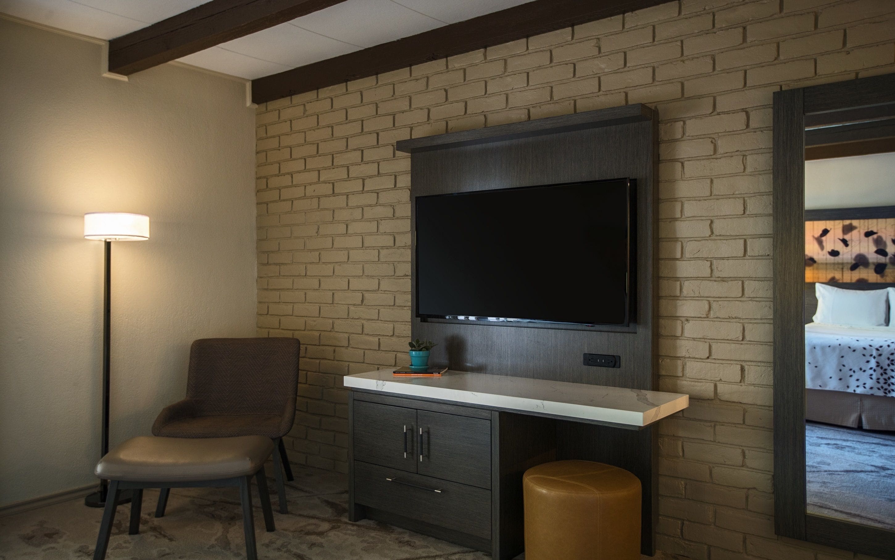 Una habitación con pared de ladrillo, un televisor de pantalla plana sobre un escritorio, una silla y una pequeña planta en maceta.