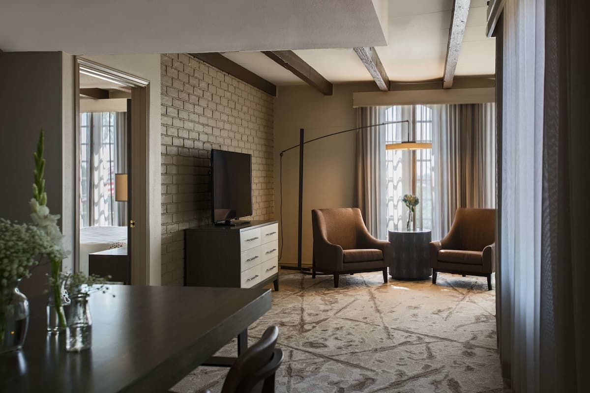 Habitación de hotel con pared de ladrillo, televisor de pantalla plana, dos sillones, una mesa redonda y un ventanal con cortinas.