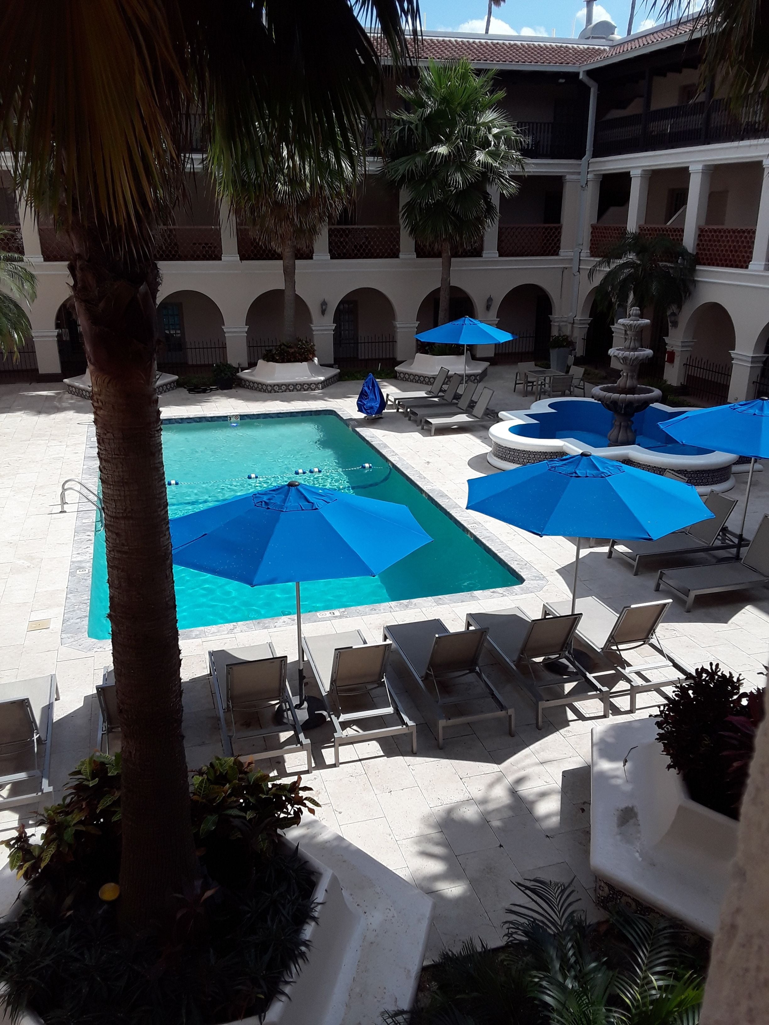 Un área de piscina con sillones de descanso y sombrillas, rodeada de palmeras y un edificio con arcos.