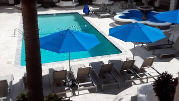 Un área de piscina con sillones de descanso y sombrillas, rodeada de palmeras y un edificio con arcos.