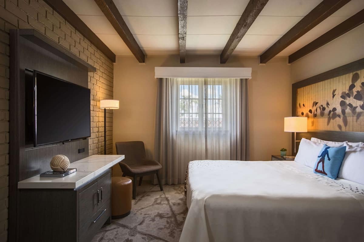 Una habitación de hotel con televisión de pantalla plana, una cama con ropa blanca, una silla, una mesita y una ventana con cortinas.