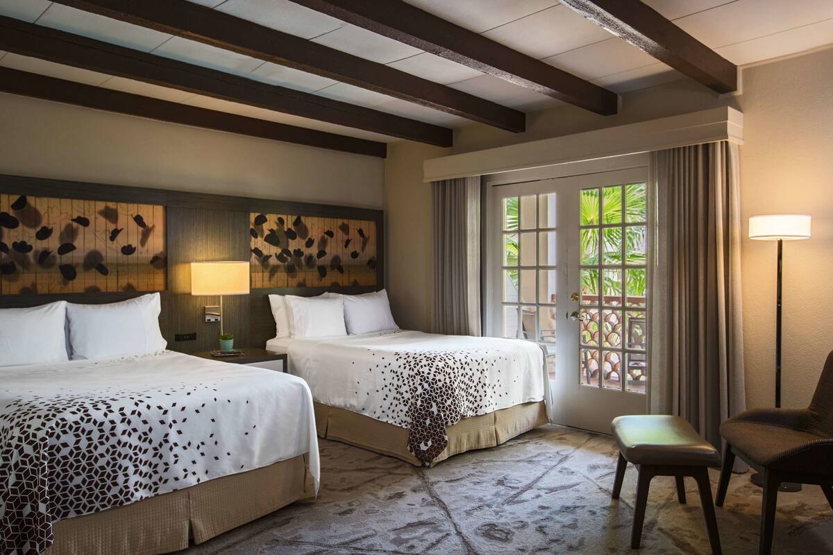 Habitación de hotel con dos camas, un banco, una silla y un ventanal con cortinas.