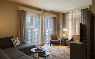 Una sala de estar moderna con un sofá, sillones, una mesa de centro y un televisor. Amplios ventanales con cortinas translúcidas dejan entrar la luz natural.
