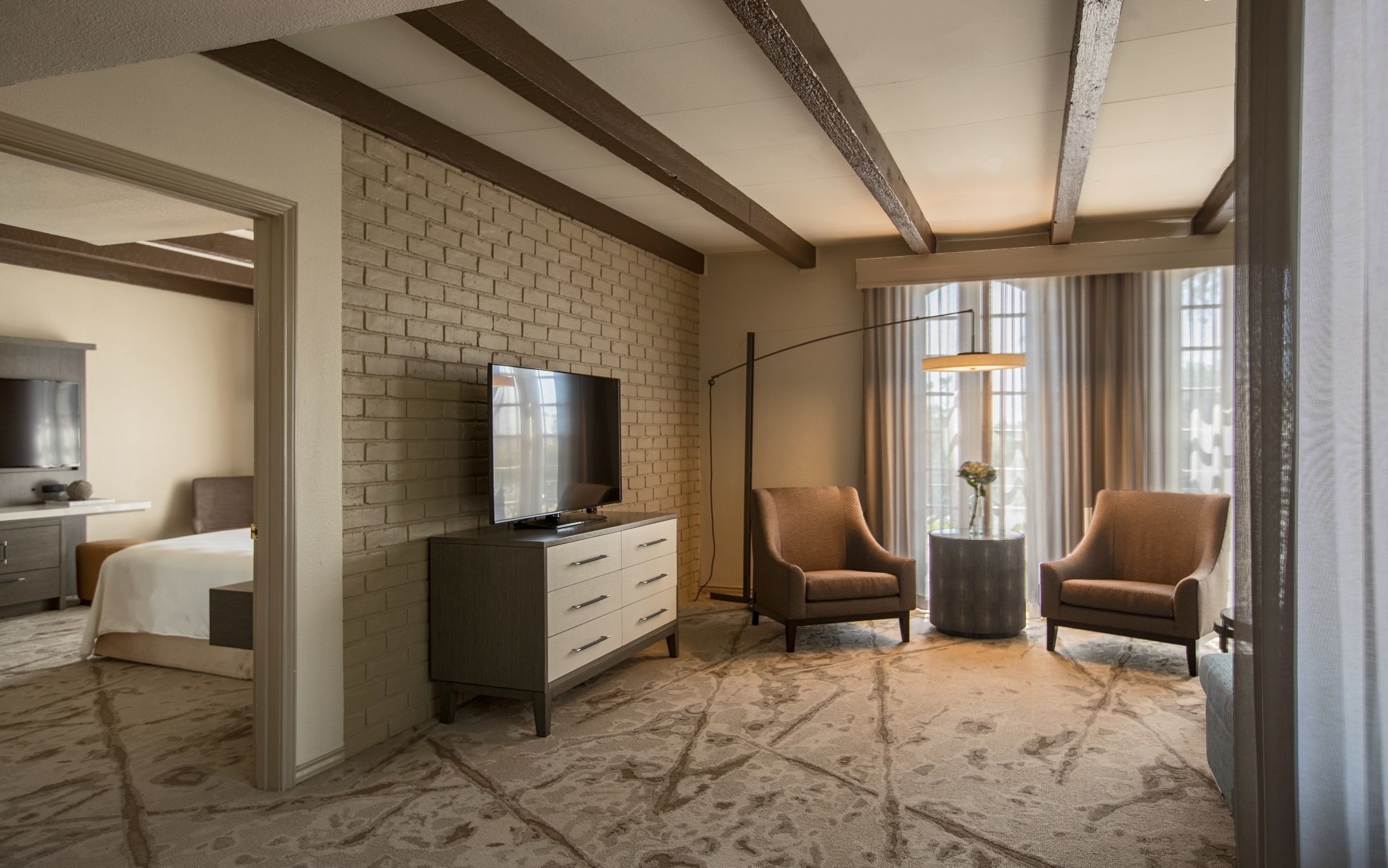 Habitación de hotel con pared de ladrillo decorativa, un televisor de pantalla plana, dos sillones, una mesita auxiliar con un jarrón y un ventanal con cortinas.