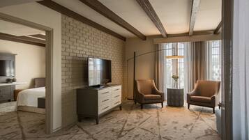 Habitación de hotel con pared de ladrillo decorativa, un televisor de pantalla plana, dos sillones, una mesita auxiliar con un jarrón y un ventanal con cortinas.