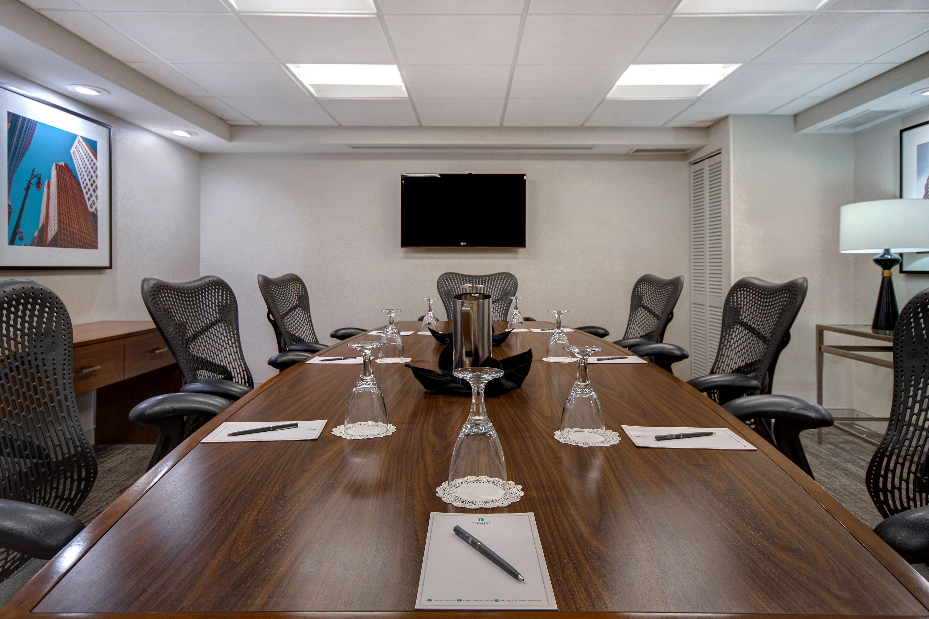 A conference room with a long wooden table, black mesh chairs, and a flat-screen TV on the wall.