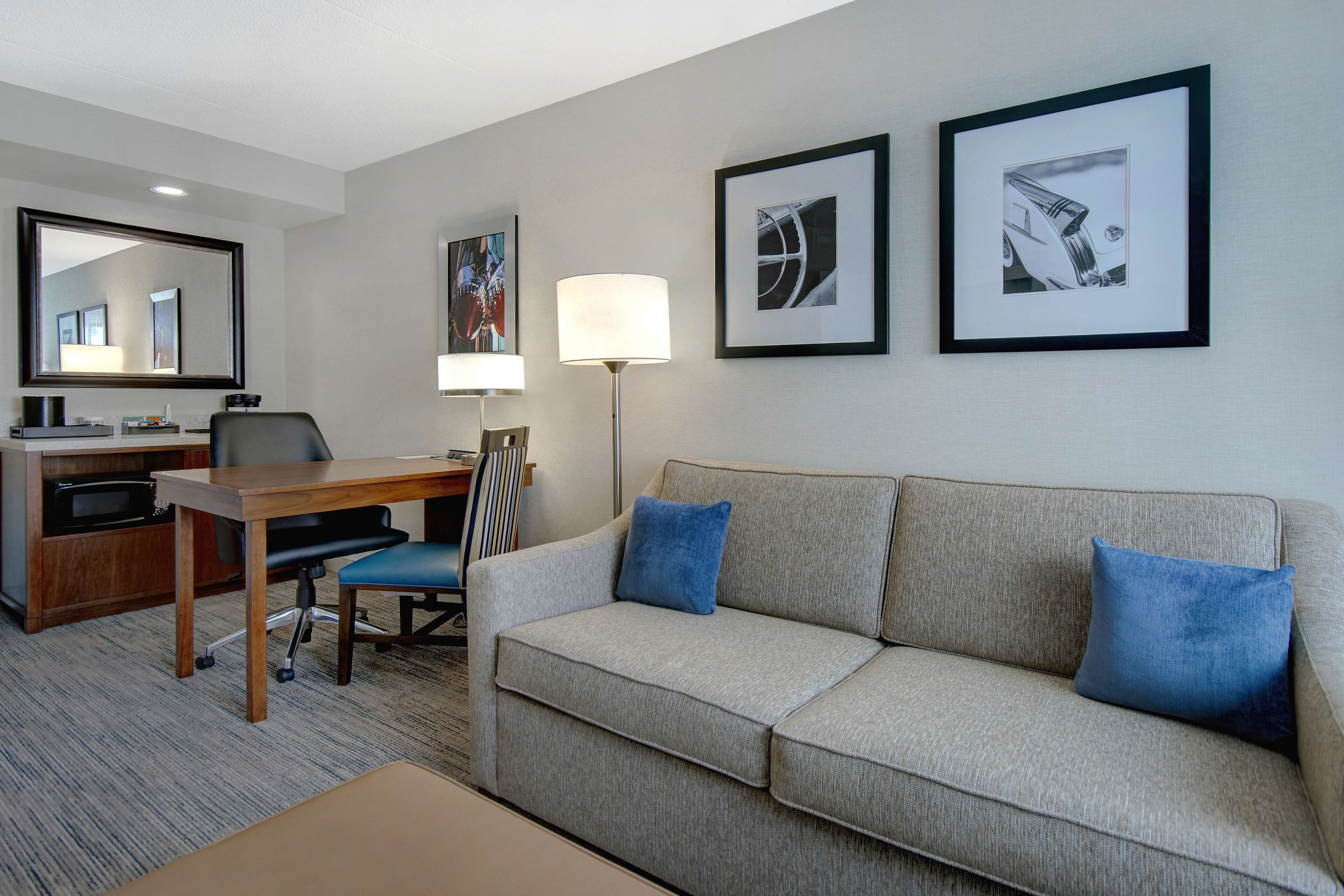 Ein Hotelzimmer mit grauem Sofa, zwei blauen Kissen, einem Schreibtisch mit Stuhl, einer Lampe und gerahmten Kunstwerken an der Wand.