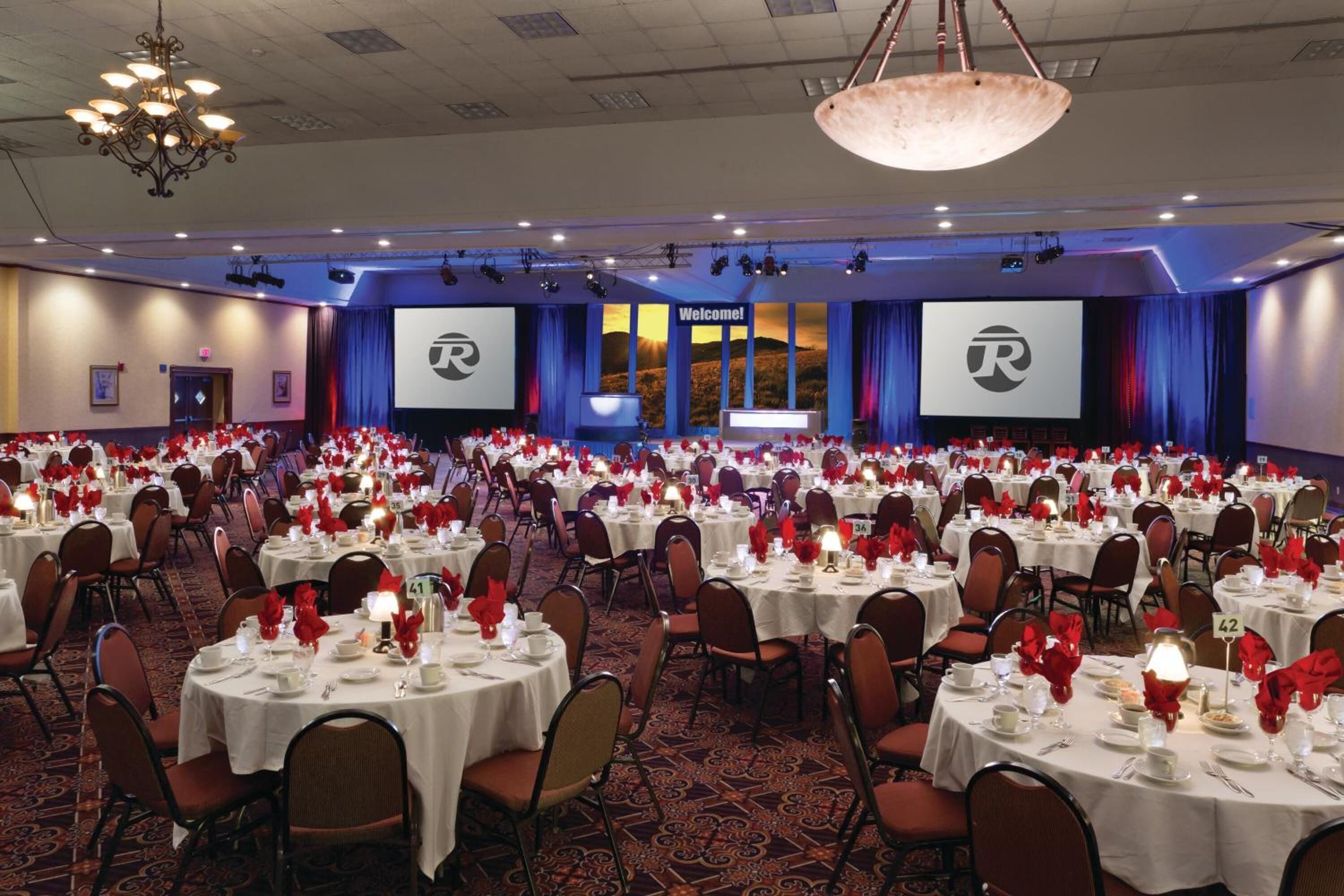 Un salón de banquetes con mesas redondas dispuestas para un evento formal, que incluye una pantalla grande y un letrero con la palabra 'Welcome!'.
