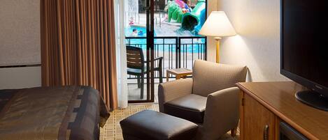 A hotel room with a bed, a TV, a desk, a chair, an ottoman, and a view of an indoor pool area.