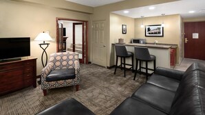 A hotel room with a sofa, a TV, a lamp, a patterned chair, a kitchenette, and a door leading to a bathroom.