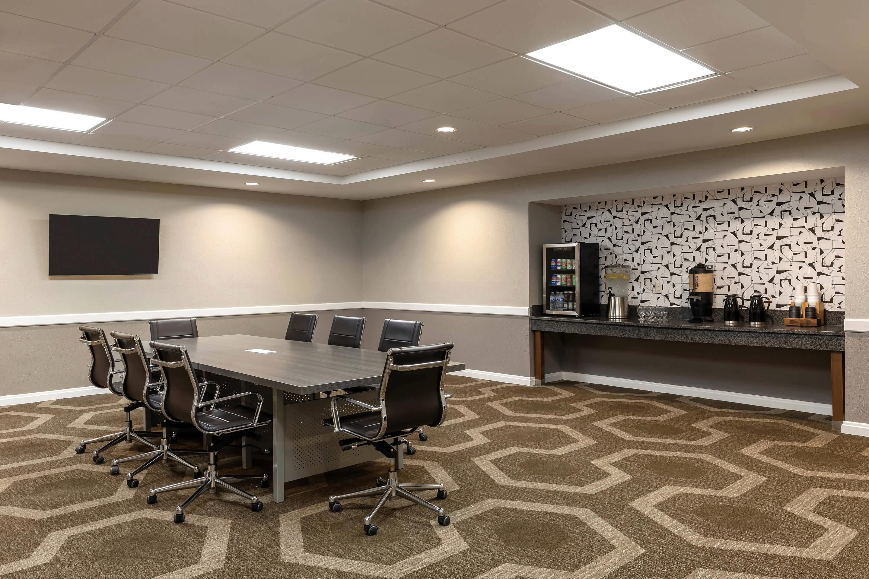 A conference room with a long table, six chairs, a coffee station, and a patterned carpet.