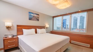 Habitación de hotel con una cama grande, cabecera de madera, dos mesitas de noche con lámparas, un cuadro en la pared y vistas de edificios altos a través de amplios ventanales.