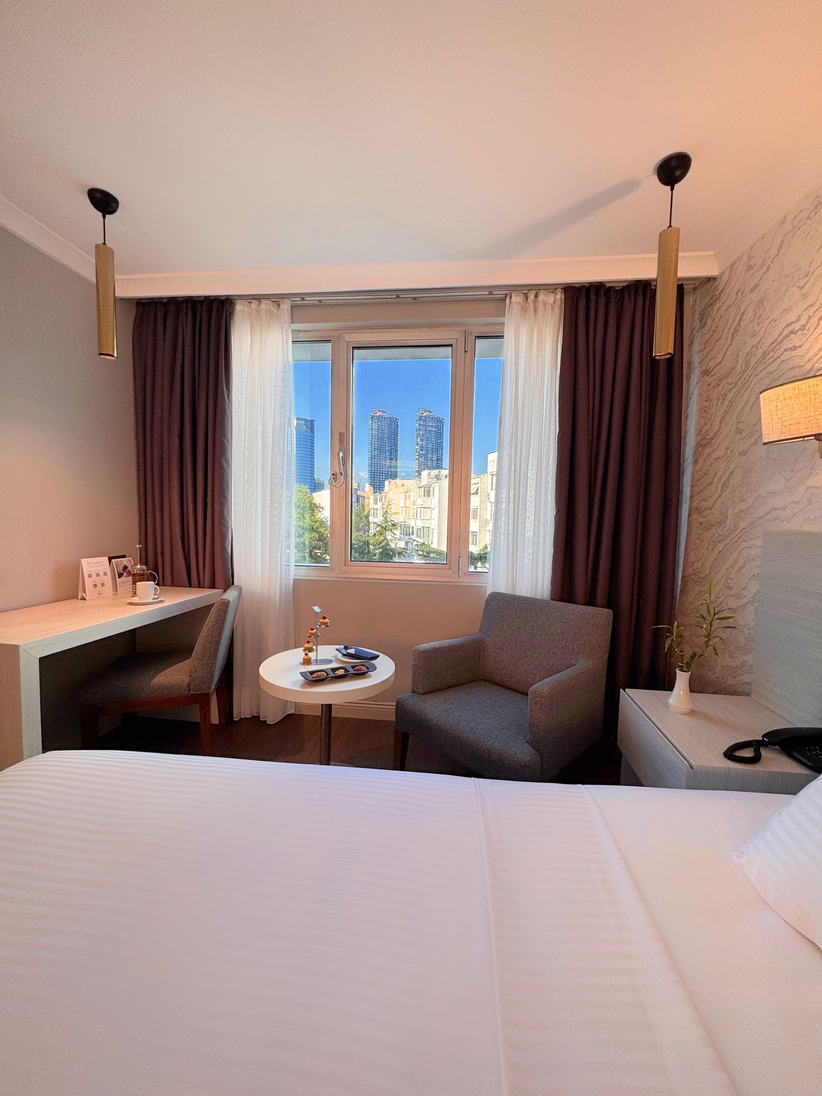 A hotel room with a bed, a desk, a chair, a small table with a tray of cookies, a vase with a plant, and a view of buildings through the window.