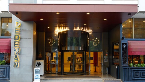 Entrada de un hotel con puerta de cristal, un letrero que dice "DEDEMAN" y un pequeño cartel con texto.