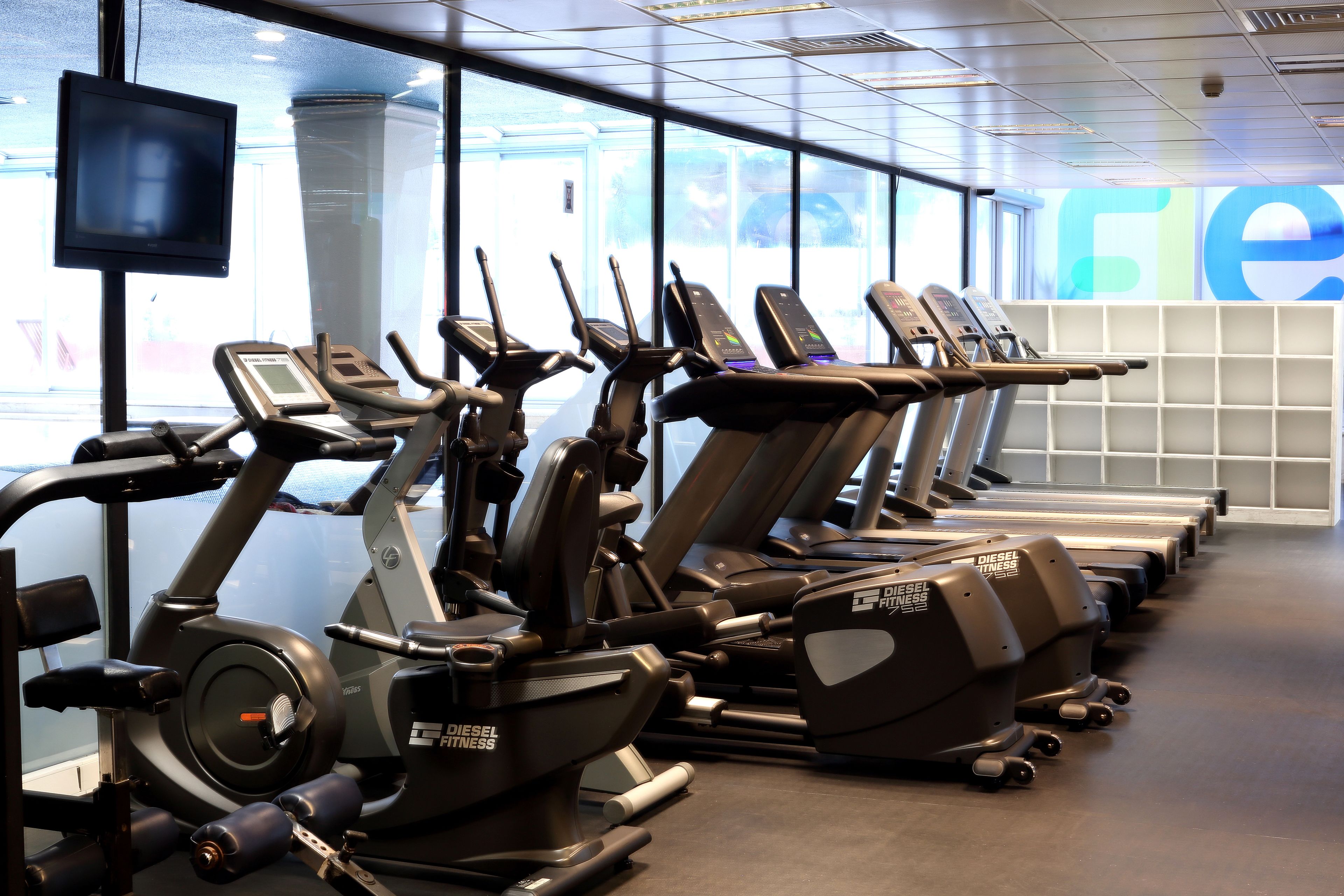 A row of exercise machines in a gym with a large window and a television mounted on the wall.