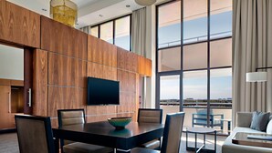 A modern hotel room with a large dining table, comfortable seating, a flat-screen TV, and large windows offering a view of the cityscape. - San Antonio Marriott Northwest Medical Center (San Antonio)