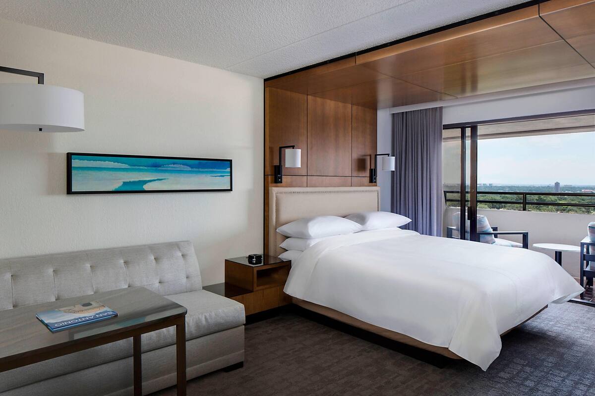 Habitación de hotel con cama, sofá, mesita pequeña, mesita de noche, un cuadro y balcón con vistas.