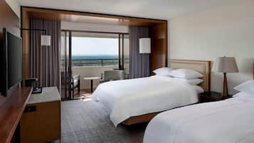 A hotel room with two beds, a desk, a TV, and a view of the city.