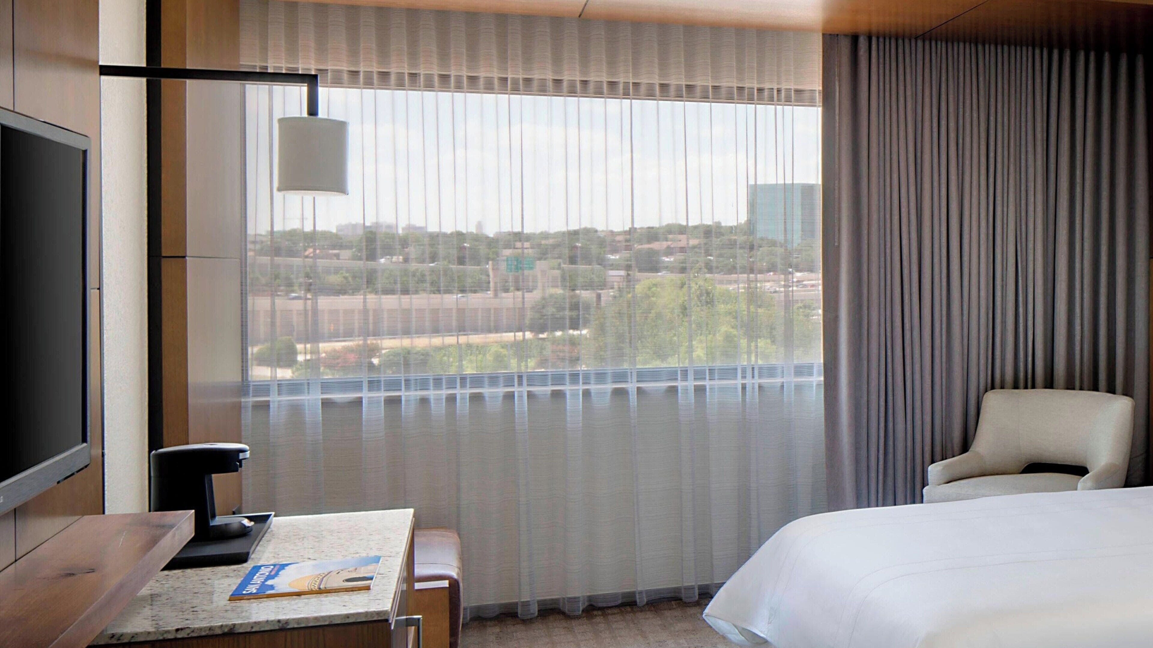 Habitación de hotel con cama, escritorio con lámpara, una silla y vista de un paisaje urbano a través de cortinas traslúcidas.