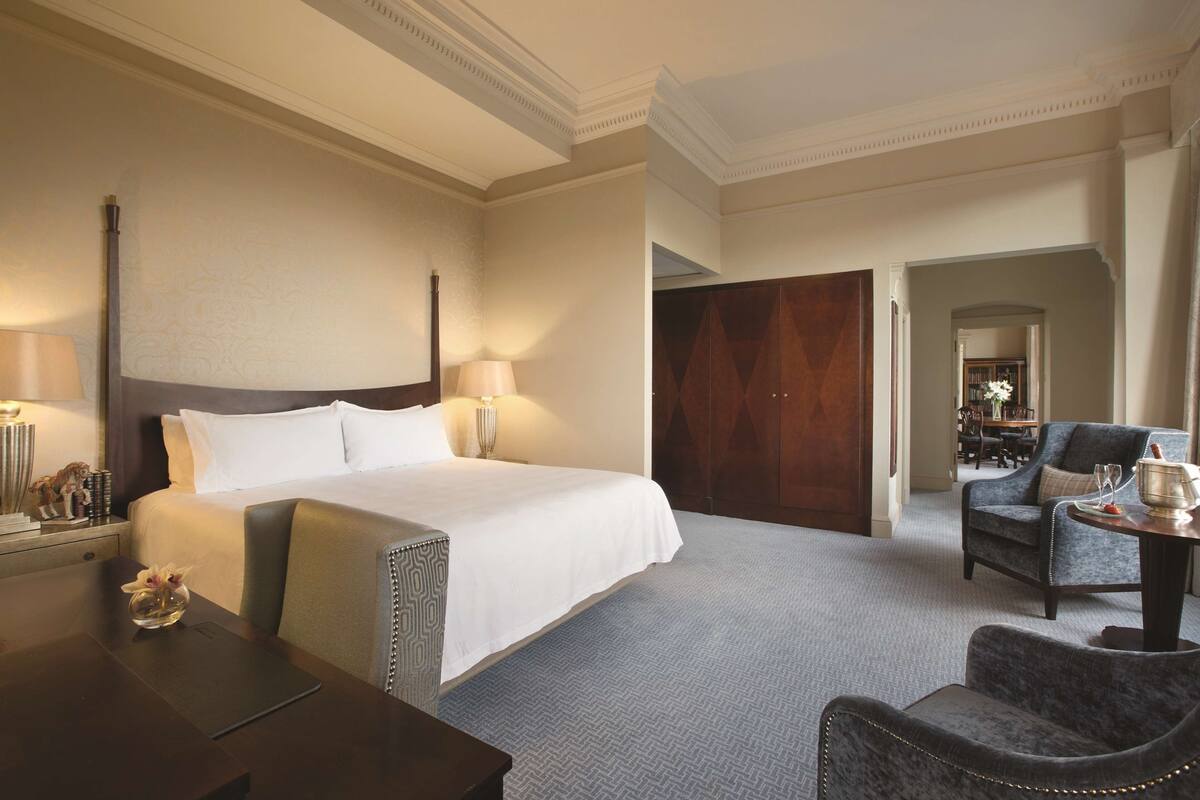 A hotel room with a large bed, a desk with a lamp, a chair, and a view of a dining area.