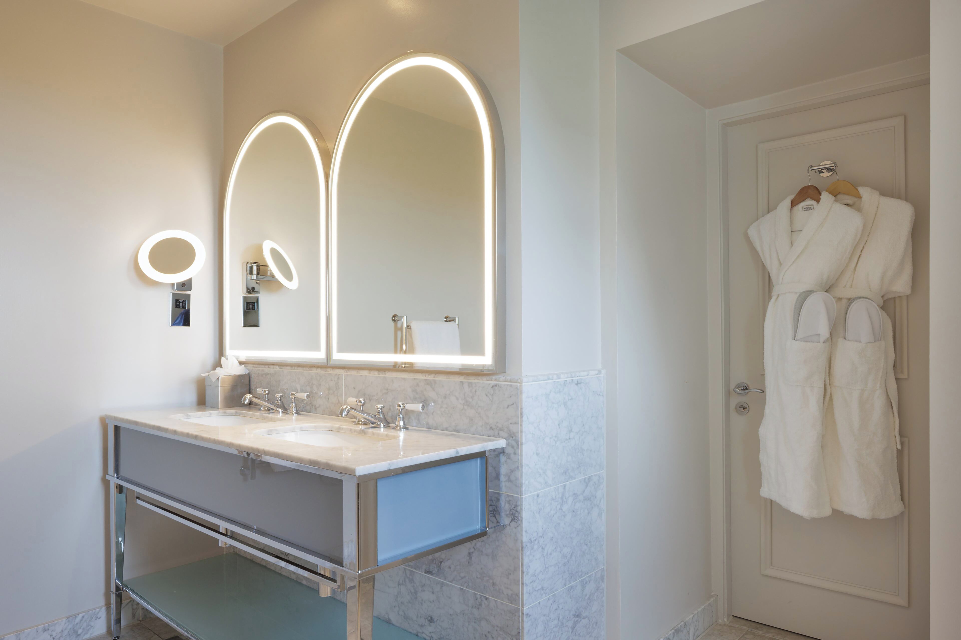 A bathroom with a double sink vanity, a large mirror with integrated lighting, and a door with a robe hanging on a hook.