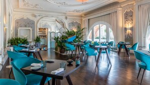 A spacious dining area with high ceilings, ornate moldings, and a chandelier. Tables are set with blue chairs and potted plants. - The Caledonian Edinburgh, Curio Collection by Hilton (Edinburgh)